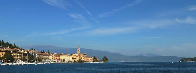 Trip Gutschein beliebte Urlaubsziele am Gardasee -  Mit einer Fläche von 370 km² ist der Gardasee der größte See Italiens. Es liegt am Fuße der Alpen und erstreckt sich über drei Staaten: Lombardei, Venetien und Trentino. Die maximale Tiefe des Sees beträgt 346 m, er hat eine längliche Form und sein nördliches Ende ist sehr schmal. Dort ist der See von den Bergen der Gruppo di Baldo umgeben. Du trittst aus deinem gemütlichen Hotelzimmer und es begrüßt dich die warme italienische Sonne. Du blickst auf den atemberaubenden Gardasee, der in zahlreichen Blautönen schimmert - von tiefem Dunkelblau bis zu funkelndem Türkis. Majestätische Berge umgeben dich, während die Brise sanft deine Haut streichelt und der Duft von blühenden Zitronenbäumen deine Nase kitzelt. Du schlenderst die malerischen, engen Gassen entlang, vorbei an farbenfrohen, blumengeschmückten Häusern. Vereinzelt unterbricht das fröhliche Lachen der Einheimischen die friedvolle Stille. Du fühlst dich wie in einem Traum, der nicht enden will. Jeder Schritt führt dich zu neuen Entdeckungen und Abenteuern. Du probierst die köstliche italienische Küche mit ihren frischen Zutaten und verführerischen Aromen. Die Sonne geht langsam unter und taucht den Himmel in ein leuchtendes Orange-rot - ein spektakulärer Anblick.